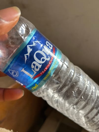 The image captures a clear, ribbed plastic water bottle held by a person's hand. The bottle shows signs of condensation or water droplets on its surface. A blue and white label wraps around the bottle, prominently featuring the brand name "LaQua" in white script. Above the brand name, a red banner reads "PREMIUM WATER" and "QUALITY YOU CAN TRUST". Below the main logo, a circular icon indicating "pH BALANCED" is visible, along with "75cl" for the volume.

The hand holding the bottle has a medium to dark skin tone, with a fingernail visible on the finger gripping the bottle's base. The background is out of focus, displaying muted, light brown or beige tones, suggesting an indoor environment. No specific time of day or weather can be discerned. The activity taking place is simply a hand holding a water bottle, which appears to be partially used. Given the location context of Ashipa, Nigeria, LaQua is likely a regional bottled water brand.