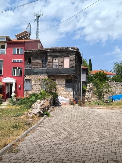This image depicts a street scene in Eğirdir, Turkey, featuring a stark contrast between an old, dilapidated wooden and stone building and a modern, red multi-story structure. The old building, situated to the right, shows significant decay with weathered wooden planks, broken windows, and a crumbling stone foundation. In front of it, a patch of dry grass and scattered rocks are visible. Adjacent to this, a modern red building with clean white-framed windows and a white awning over an entrance stands prominently. A cell tower and a solar water heater are visible on the rooftop of the red building, indicating a more contemporary infrastructure. The foreground is dominated by a paved cobblestone walkway or street. The background reveals lush green trees and other buildings with terracotta roofs, suggesting a residential or mixed-use area. The sky is partly cloudy with blue patches. No people are present in the frame, and no actions or interactions are depicted. The scene appears to be an outdoor, daytime setting.