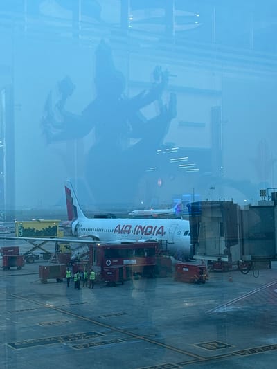An Air India plane is at an airport gate, viewed through a window, with reflections of indoor structures. The plane is white with red Air India lettering. Airport personnel surround the plane. Ground support equipment, including baggage handling vehicles, is connected or nearby. The gate area has visible markings on the pavement. The window distorts the view with superimposed reflections of shelves and other interior elements. The overall scene is muted in tone.