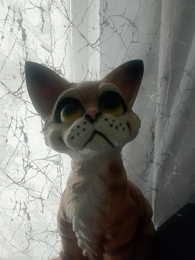 A clay cat figurine with large, expressive eyes and pointed ears is positioned in front of a sheer curtain with a marbled pattern. The figurine's fur is textured and painted in shades of brown and tan, with black dots on its muzzle. The cat is looking upwards with a slightly mournful expression. The background consists of a white curtain with dark, web-like lines, and a hint of a dark object to the right of the figurine. The overall lighting is soft and diffused, suggesting an indoor setting. There is no indication of the location being in Berdyaush, Russia.