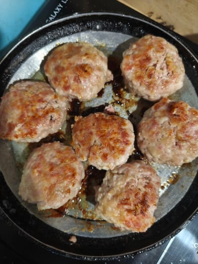 This is a close-up, overhead shot of seven round, uncooked meat patties in a dark frying pan. The patties are light brown with some pinkish meat visible, and appear to be made of ground meat, possibly pork or beef. They are arranged in a circular pattern within the pan, with some dark brown drippings and grease pooling around them. The pan itself is round and appears to be made of metal, with a black rim. In the background, a black surface with "GALAXY L" printed in white text is visible. The lighting suggests an indoor setting, likely a kitchen. The overall impression is of food being prepared for cooking.