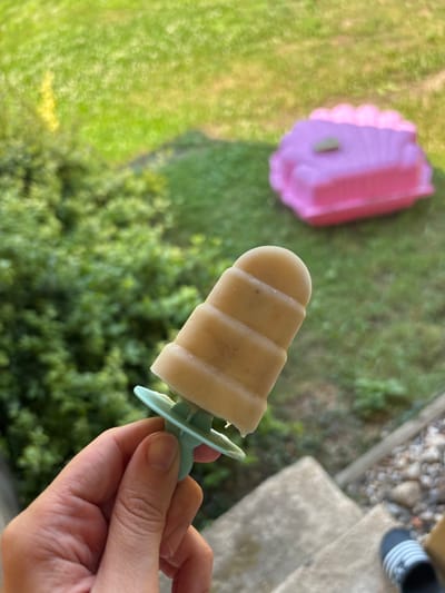 A hand holds a light brown popsicle with a light blue plastic handle. The popsicle has horizontal ridges. The background is out of focus, showing green grass and trees, suggesting an outdoor setting. To the left, a pink plastic mold is visible, likely used to create the popsicle. In the upper right, there are concrete steps and a person's foot wearing a black and white striped sandal. The lighting suggests it is daytime, possibly sunny, given the bright green of the grass and the shadows. There are no visible text elements in the image. The scene conveys a sense of casual outdoor activity, possibly related to making or enjoying homemade frozen treats on a warm day.