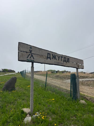 The image depicts a weathered wooden sign indicating "Джугди Глэмпинг" (Jugdi Glamping), likely an advertisement for a glamping site. The sign is made of two planks mounted on posts wrapped in rope. Below the text are symbols resembling a diamond shape, crossed sticks, and a wavy line. The sign stands beside a chain-link fence in a grassy area with yellow wildflowers. A dirt road runs parallel to the fence. In the background, there are buildings, possibly part of a village or campsite. The image suggests a rural setting, and the location is Zarubino, Russia. No people are visible.