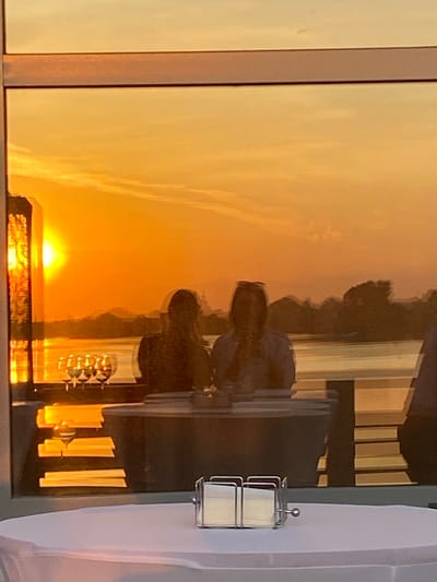 The image shows a window reflecting a vibrant sunset over water, likely a lake or river. The reflection also captures silhouettes of trees or buildings along the horizon, and structures closer to the viewer, possibly part of a building or indoor decor. An object is attached to the wall near the window with a silver stand. There is no information about the location in the city of Aramil, Russia.