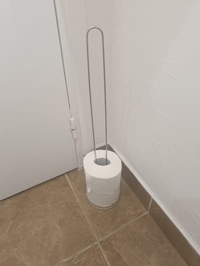 A toilet paper holder stands on a tiled floor beside a white door and wall. The holder is a simple, silver-colored frame with a tall, looped handle. The toilet paper is stacked high, nearly reaching the handle's base. The tiles are a warm, brown color with subtle veining. The wall has a slightly textured, painted finish. The overall scene is a clean, functional bathroom space.
