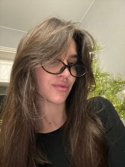 A female individual is depicted in an indoor setting, identified as the city of Rasskazovo, Russia. The subject occupies the majority of the frame, facing slightly downwards and to the viewer's right. She has long, layered brown hair with bangs that partially obscure her forehead. She is wearing dark, tortoise-shell rimmed eyeglasses. Her lips are slightly parted. A small, dark mole is visible on her upper neck, below her jawline, on the viewer's left side. She is attired in a dark, possibly black, crew-neck garment. In the background to the left, a light-colored wall with white molding is partially visible. To the right of the subject, a blurred green object, consistent with a pine or fir branch, is present, decorated with numerous small, illuminated white lights and at least one spherical white ornament. The lighting suggests an indoor environment.