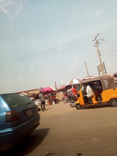 The image depicts a dynamic street scene, likely in Asokoro, Nigeria, under a bright, clear daytime sky. The setting appears to be a bustling roadside area with a mix of commercial activity and vehicle traffic.
In the foreground on the left, the rear of a blue car is partially visible, suggesting movement on the dusty road. To the right, a prominent yellow tricycle, known locally as a keke napep, is seen with passengers inside, indicating public transport activity. Adjacent to it, a motorcycle is parked near a cluster of informal stalls.
These roadside stalls are characterized by vibrant, open umbrellas, including pink and brown-and-white striped ones, providing shade for vendors and their goods. A person in a striped shirt stands near one of these stalls. In the background, a large red metal container, possibly repurposed as a shop, adds to the commercial landscape. Visible on one of the stalls is the number "200", which likely denotes a price.
Overhead, multiple utility poles support a network of power lines, typical of an urban or semi-urban environment. The overall atmosphere conveys a sense of daily commerce and movement in a lively community setting.