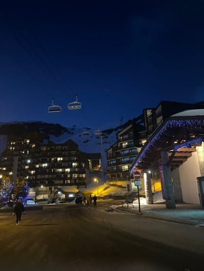 The image depicts a nighttime scene in a snowy mountain resort within Saint-Martin-de-Belleville, France. The deep blue sky, indicative of dusk or early morning, features a single faint celestial body in the upper right quadrant. Overhead, multiple ski lift chairlifts are visible suspended by cables, with at least two prominent chairs in the mid-ground.
The environment is characterized by snow-covered ground and mountains in the background. Several multi-story chalet-style buildings line the scene, featuring numerous illuminated windows, suggesting occupancy. On the far left, a decorated tree adorned with blue lights is present.
In the foreground, a single individual wearing dark clothing walks towards the viewer on a cleared, possibly wet or icy, paved surface. Further back, near the center of the image, a group of approximately four to five individuals stands conversing on the snow-covered ground near a smaller building entrance.
To the right, a large building with a prominent wooden entrance overhang is illuminated by blue string lights. Adjacent to this structure, a "No Entry" sign (red circle with white horizontal bar) is visible on a pole, with a small dark bench situated nearby. Streetlights provide yellow and white illumination to various areas of the scene, casting shadows.