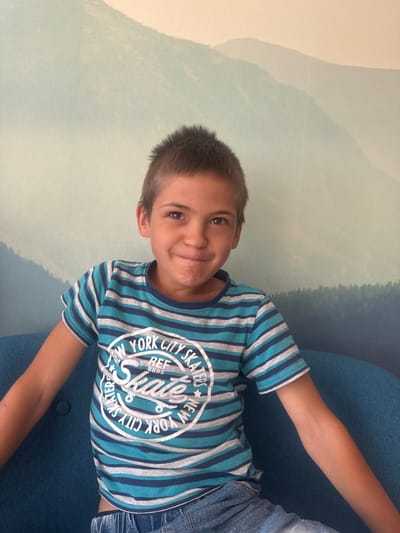 A young boy with short, spiky brown hair and brown eyes is seated on a blue cushioned bench. He is wearing a striped t-shirt with horizontal bands of blue and grey, and blue denim shorts. The t-shirt has a circular graphic on the front that says "NEW YORK CITY SKATERS" with "Skate" in the center. The boy is looking at the camera with a slight smile. The background is a mural of a hazy, mountainous landscape with trees. The image appears to be taken indoors, possibly in a public space or a home. No specific location in Divonne-les-Bains, France, can be identified from the image itself.