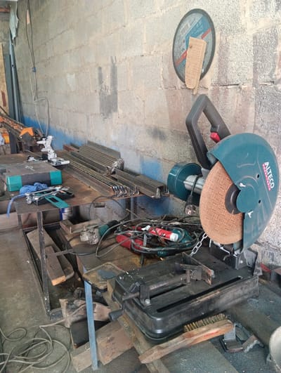 The image shows a workshop in Granovshchina, Russia. A large chop saw with a teal guard and an ALTECO POWERTOOLS brand is prominently displayed. A pile of L-shaped metal bars is arranged neatly on a metal shelf. Various tools, including wrenches and a blue cloth, are on a workbench. A small angle grinder, red and black electrical cords, and a chain are also visible on the workbench. In the background, a concrete block wall is visible, with a circular saw blade attached to it with a piece of cardboard. The floor appears to be concrete and is cluttered with electrical cords and wooden planks.