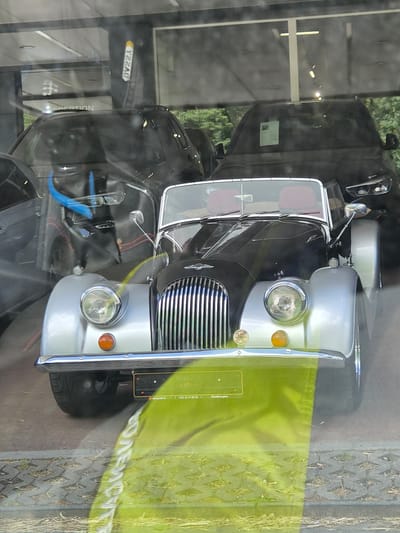 The image shows a vintage silver and black convertible car, likely a Morgan, parked behind a glass window. The car has a chrome grille and red interior seats. In the background, two dark-colored SUVs are visible. A bright green fabric is draped over the front of the Morgan. A license plate frame with text, including "+352 26 37 60 00 | TotalEnergies" is attached to the front of the car. The background appears to be a car dealership in Bettembourg, Luxembourg, with trees visible outside the building. Reflections on the glass suggest the image was taken from outside.