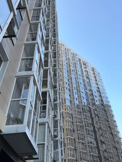 The image shows a modern building with a stepped facade, covered in numerous windows. The windows on the upper floors are largely open, suggesting ventilation or a particular architectural feature. The lower part of the building features a series of glass-enclosed, box-like structures that appear to be extensions or balconies, also with reflective surfaces. The sky is clear and blue, indicating daytime with fair weather. There are no people visible in the image. The architecture is geometric and repetitive, with a focus on glass and metal. The scene is likely an exterior shot of a contemporary building, possibly an office or residential complex, viewed from a slightly upward angle. The location context provided (Elizavetinskaya, Russia) is not evident from visual cues in the image itself. No text is visible in the image.