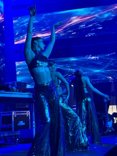 In Akhalubani, Georgia, three women in shimmering, sequined outfits perform on a stage illuminated by vibrant blue lights. The woman in the foreground, facing left with her back to the viewer, has her arms raised and her body angled, showcasing a sequined halter top and wide-leg pants. To her left, another woman, facing the same direction but partially obscured, is also in similar attire. The third woman, on the far right, faces away from the camera, her long dark hair flowing as she performs with her arms extended. Behind them, a large screen displays a dynamic, abstract pattern of blue and pink light, creating an ethereal backdrop. On the left side of the stage, a DJ setup is visible, with equipment and a person's hands operating it. Stage lighting and sound equipment, including speakers and a guitar on a stand, are present in the background, suggesting a musical performance. The overall atmosphere is one of energetic entertainment.
