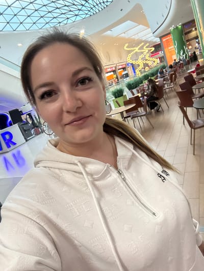The image is a selfie taken by a woman inside a modern shopping mall. The woman, with light brown hair pulled back, is wearing a white zippered hoodie. She is positioned in the foreground, her face filling the upper portion of the frame. In the background, the mall's interior is visible, characterized by a domed glass ceiling, rows of shops, and a food court area with tables and chairs. A large decorative light fixture, shaped like a chain of yellow circles, hangs from the ceiling in the middle ground. People are seated at tables in the food court, engaged in conversations or eating. A sign with Cyrillic text is visible on one of the shops. The flooring appears to be tiled. The lighting suggests it is daytime, with natural light filtering through the ceiling. The location is identified as Tlustenkhabl, Russia.