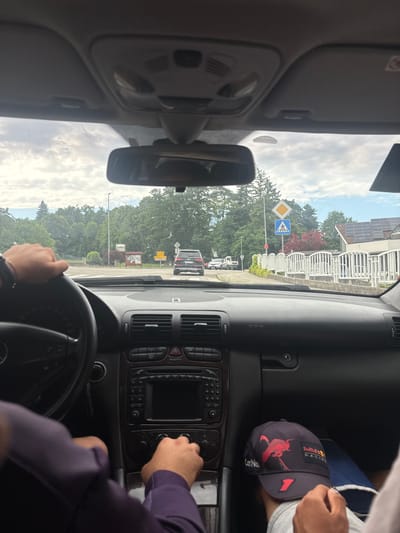 The image depicts the interior of a car in Nova vas pri Mokricah, Slovenia, viewed from the perspective of the driver. A person's hands are visible, one on the steering wheel and the other operating the car's central console. Outside the car, a black SUV is seen driving ahead on a road lined with trees and buildings. Traffic signs, including a pedestrian crossing sign, are also visible. The sky is overcast with clouds. The car interior shows a dashboard with the car's radio and air conditioning controls. A baseball cap with the Red Bull Racing logo is placed on the dashboard.