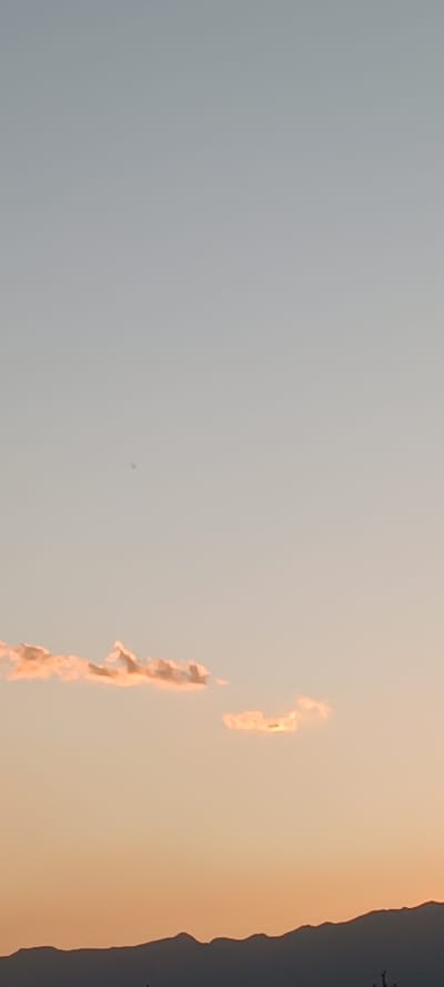 The image shows a panoramic view of the sky at dusk with a silhouetted mountain range in the foreground. The sky transitions from a pale blue at the top to a soft orange and peach hue near the horizon. Several wispy clouds, tinged with pink and orange from the setting sun, are scattered across the sky. A small, dark speck is visible in the upper portion of the sky, possibly a bird or insect. The mountain range is dark and undulating, suggesting a natural landscape. The overall scene evokes a sense of tranquility and the beauty of nature at sunset. The location is identified as Chyrpykty, Kyrgyzstan.
