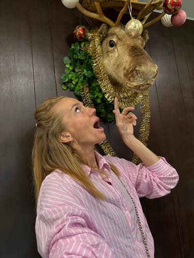In Bolotnitsa, Russia, a woman with blonde hair, wearing a pink and white striped shirt and a silver chain necklace, stands in front of a dark wooden wall. She is looking up and to her right, with her mouth open, as if surprised or excited. Her right hand is raised, with her index finger pointing upwards. Above her, a mounted reindeer head is decorated with a green wreath, gold tinsel, and several Christmas ornaments, including a red and gold bauble, a textured white ornament, and a shimmering gold bauble. The scene appears to be indoors, possibly in a home or establishment decorated for the holiday season.