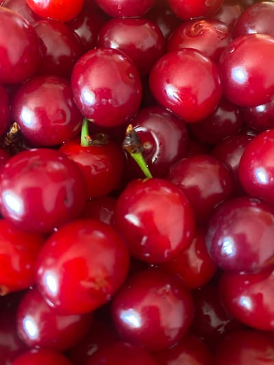 This is a close-up, overhead shot of a pile of ripe, red cherries. The cherries are plump and glossy, with a deep red hue and some variations in shade. The lighting suggests a bright environment, possibly natural daylight, as evidenced by the reflections and highlights on the fruit's surface. There are no people or text visible in the image. The setting appears to be a domestic or agricultural context, focusing entirely on the cherries. The image captures the texture and rich color of the fruit, emphasizing its freshness and abundance. The composition is tightly cropped, filling the frame with the cherries, suggesting a focus on the subject itself rather than its surroundings.