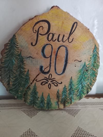 The image shows a wooden slice decorated with painted trees, the name "Paul", and the number "90". The background is painted yellow, suggesting a sunset or sunrise. The wood slice is placed on a patterned tablecloth. There are no people or animals visible in the image. The location in the city of Hohentengen, Germany is not specified in the image or its context.