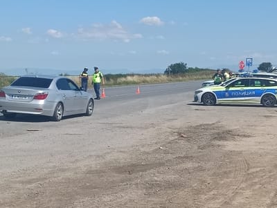 The image depicts a roadside scene in Tenevo, Bulgaria, on a clear, sunny day. Two police officers are present, one in a yellow high-visibility vest and the other in a dark uniform with a high-visibility vest. They are standing near orange traffic cones on the asphalt road. A silver BMW sedan is parked on the left side of the road.
Further down the road, two police cars are visible, one white and blue with "ПОЛИЦИЯ" (Police) written on it, and another partly visible. A stop sign is mounted on a pole behind the police cars. The background consists of a grassy field with distant hills and a bright blue sky with scattered clouds. The overall impression is of a police checkpoint or a traffic stop operation.
