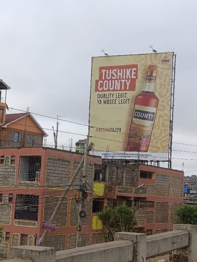 An outdoor billboard promotes Tushike County liquor. The bottle's label reads "County," and the slogan states, "Quality Legit, Ya Wasee Legit." A hashtag, #KESHOITAJUPA, is included. The billboard also contains a health warning regarding alcohol consumption and prohibits sale to individuals under 18. The billboard is positioned above a building in a densely populated area.