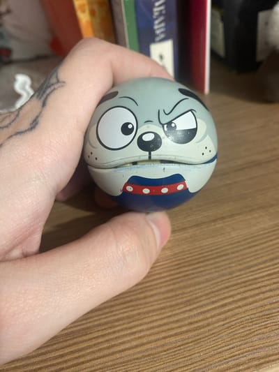 A close-up, slightly tilted shot shows a person's hand holding a light blue, spherical toy with a cartoon dog face. The dog has large, white eyes with black pupils, an expressive eyebrow, and black spots. It appears to be wearing a red collar with white dots, bordered by a blue stripe. The toy has a seam running vertically along its face, suggesting it might be a container or two halves that fit together. The background is a warm, wooden surface with distinct vertical grain. To the left of the toy, out of focus, are parts of what appear to be boxes or packaging, with some text visible on the blue packaging that reads "АКАЦИЯ". The lighting is neutral, and the scene appears to be indoors, likely in a home or office setting, given the wooden surface and the presence of packaged goods. The image doesn't convey a specific time of day or weather conditions. The focus is on the toy and the hand, with a hint of a tattoo visible on the person's wrist.