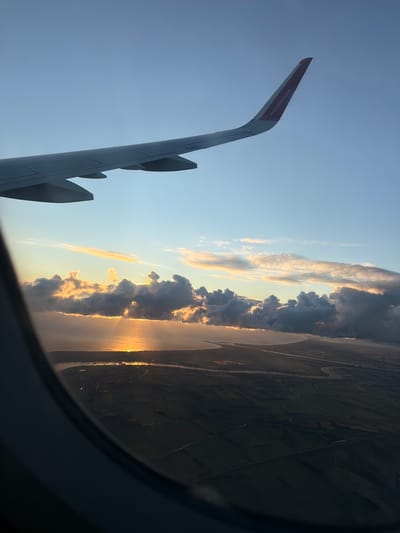 An aerial view captured from an airplane window shows a landscape at sunset. The upper part of the image is dominated by a clear blue sky with a segment of an airplane wing extending from the left, featuring the Wizz Air logo on its tip. Below the wing, a horizon line separates the sky from the earth. Dark clouds are illuminated from behind by the setting sun, which casts golden rays across the water and the land. The water appears to be a large body, possibly a sea or a lake, with a coastline visible. The land below the water is a patchwork of agricultural fields, with some structures and roads subtly visible. The view is framed by the dark curve of the airplane window. The location in the city of Czerwony Kościół, Poland, is suggested by the presence of a Wizz Air flight.
