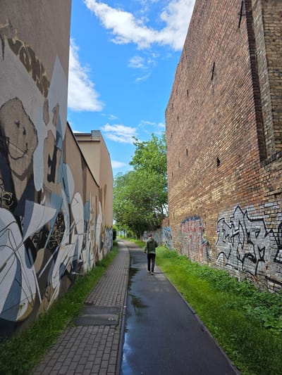In Halle (Saale), Germany, a person walks along a wet paved path between two buildings. The building on the left is adorned with a large mural. The building on the right is constructed of brick and covered in graffiti. The sky above is blue with scattered white clouds. Green trees are visible in the background.