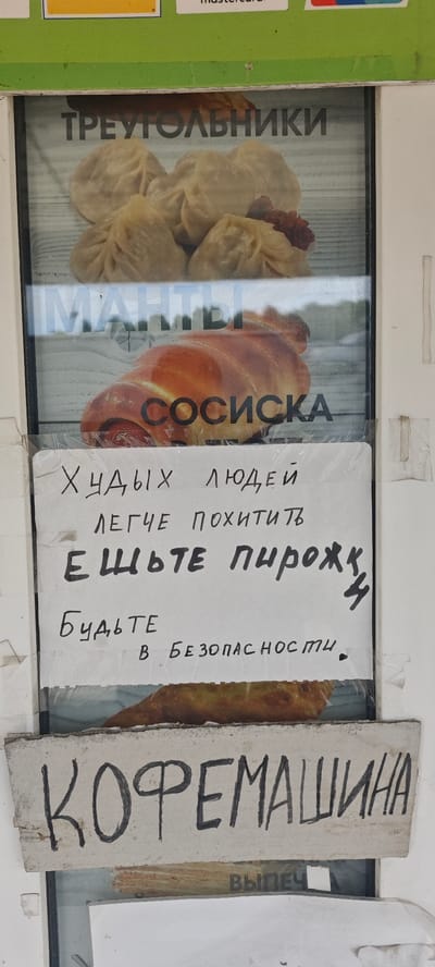 The image shows a window display in Nizhnie Yaki, Russia. The top of the window has a MasterCard logo. Inside the window are advertisements for food items. At the top is a picture of what appears to be dumplings labeled "TRIANGLES". Below that is a picture of a sausage roll labeled "SAUSAGE". Pasted onto the window is a handwritten sign that reads "Thin people are easier to kidnap. Eat pies. Be safe". Below the sign, there is another picture of a pie. At the bottom of the window, there is a sign that says "COFFEE MACHINE". Below that sign, the word "BAKERY" is barely visible.