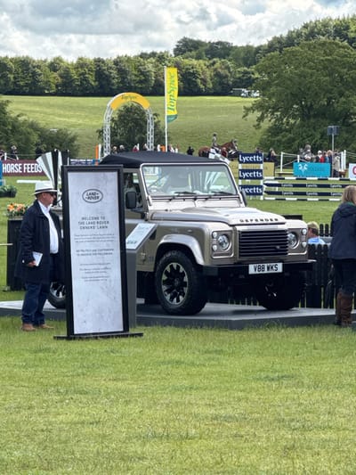 The image shows a Land Rover Defender parked on a raised platform in a grassy field at the Land Rover Owners' Lawn at the Bramham Horse Trials in Bramham, United Kingdom. A man in a black jacket and white hat is standing beside a sign that reads "Welcome to the Land Rover Owners' Lawn". The Land Rover is beige with a black top and a black license plate that reads "V88 WKS". In the background, there are a group of people standing behind a fence and a yellow "TopSpec" flag.