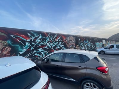 A widescreen, eye-level outdoor shot shows a graffiti-covered wall in Cartagena, Spain, under a partially clouded blue sky. The graffiti features a stylized, aggressive-looking older man with glasses and a large mustache, as well as abstract, sharp-edged lettering in teal, black, and red. Two cars are parked in front of the wall, partially obscuring the graffiti. In the foreground, a white car's rear is visible on the left, and a brown car with a white roof is parked in the center. On the right, a silver minivan is driving away, with its driver leaning out the window. The ground is paved.