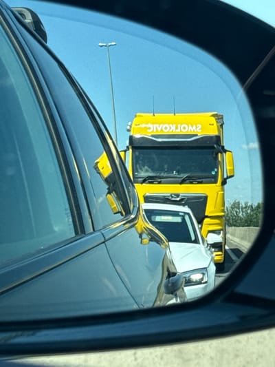 The image is a reflection in a car's side mirror, showing a yellow truck. The truck is primarily yellow with black accents. On the side of the truck, there is text that reads "2WORKOAIC" in white, which appears to be a reversed reflection of "CIAOKROW2". The truck has a white cabin. The background is a clear blue sky, suggesting daytime. There are some green trees visible in the upper right corner of the reflection. The truck seems to be stationary, and the scene is viewed from the perspective of another vehicle. The overall impression is a snapshot of traffic or a roadside view captured in a side mirror. The location is identified as Karlovac, Croatia.