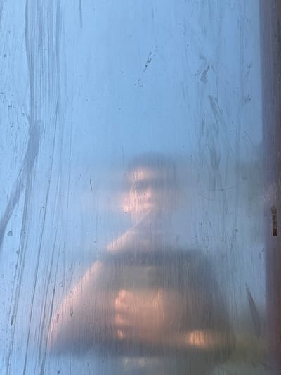 A photograph depicts a slightly blurred view through what appears to be a scratched or frosted glass surface. The overall color palette is dominated by cool tones of blue and gray. Horizontal streaks and scratches are visible across the glass, obscuring the details of what lies beyond. There's a somewhat opaque, blurry impression of what looks like possibly a person or form with a subtle light or glow. The location Longone al Segrino, Italy is not possible to confirm due to lack of recognizable landmarks and context.