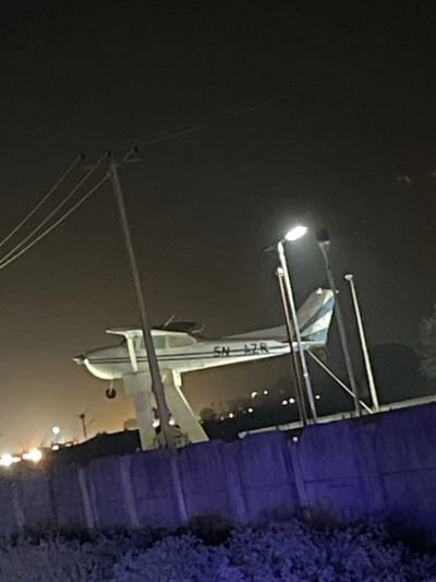An airplane with the marking "5N-4ZR" appears to be on the ground, possibly after an accident. The scene is dark, illuminated by a streetlight above the tail of the plane. Power lines are visible in the foreground. The background includes some foliage and distant lights, suggesting the location is near a developed area. The image has been rotated.