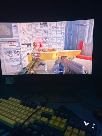 The image captures a scene featuring a computer monitor displaying a video game, positioned next to a mechanical keyboard on a dark desk. The photograph appears to be taken in a dimly lit room, with the monitor emitting the primary light. The monitor itself is shown in a portrait orientation relative to the camera's perspective, though the game content is clearly designed for a landscape display.
The monitor displays a first-person shooter (FPS) video game. The game environment features what appears to be an urban or industrial setting, characterized by light-colored, possibly concrete or brick walls arranged in a grid-like pattern. In the foreground of the game screen, a player character is visible, clad in a tan or olive military-style outfit with a balaclava covering their face. This character is engaged in combat, facing another partially visible character or target, from whom muzzle flashes and smoke are emanating, suggesting active gunfire. Red splatters, indicative of blood or damage, are visible on the ground and surrounding structures, emphasizing the intensity of the in-game action.
Several user interface (UI) elements from the game are visible:
* In the bottom left corner, a small circular minimap is present, with Russian text below it reading "Точка закладки А" (Bomb site A).
* Numbers such as "78", "73", and "76" are scattered across the game world elements, possibly marking objectives or locations.
* A health or armor bar showing "100" is located in the bottom center.
* The top left corner displays a kill feed or event log, mentioning names like "maksa" and "КИБЕРДИМАСЕТ" (KIBERDIMACET), accompanied by weapon icons, notably an AK-47.
* The bottom right corner shows weapon information, indicating an "AK-47" with an ammunition count of "28 | 90". A question mark icon is also present nearby.
* Near the top center, a score or team status section shows numbers "5" and "2" along with small flags.
To the right of the monitor, a mechanical keyboard occupies a significant portion of the desk. The keyboard has a dark base, with mostly dark keycaps, though some keycaps are a distinct lighter color, possibly yellow or light green, adding a customized aesthetic. Key labels visible include "Enter", "Ctrl", "Alt", "F10", "F12", "PrtSc", "ScrLk", "Pause", "Ins", and "Del". One key distinctly shows the text "PYSSE". On the upper right side of the keyboard, several small, glowing indicators or LEDs are visible. A mouse or a mouse cable is partially visible next to the keyboard.
The overall impression is of a gaming setup actively in use, with a player immersed in an intense combat scenario within the game. The dark environment highlights the glowing screen and keyboard, creating a typical nocturnal gaming atmosphere. A small white object, possibly an electronic device or power adapter, is visible on the desk beneath the monitor.