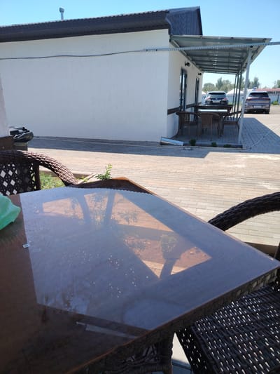 The image shows an outdoor seating area with a glass-top table and wicker chairs. The table is positioned in the foreground, reflecting the surroundings, including the sky, trees, and a building with a white textured wall and a metal roof. In the background, under a metal awning extending from the building, are more wicker chairs and tables. Two cars are parked in the distance. The ground is paved with light-colored bricks. A trash can is visible on the left side of the image, near the white building. The scene appears to be in a location with clear blue skies and bright sunlight, casting shadows. The area seems to be a cafe or a rest stop, likely in a town or rural area given the open space and parked vehicles. The city of Тораңғылық is not visible in the image, but the scene depicts an outdoor setting that could be found in Kazakhstan.