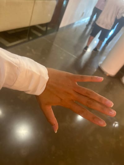 A light-skinned right hand and forearm, partially covered by a white long-sleeved garment, are prominently displayed across a dark, highly reflective polished floor surface. The hand is open, palm facing downwards, with fingers slightly spread. In the background reflection on the floor, two human figures are visible; one appears to be wearing a light top, dark shorts, and dark shoes, while the other's attire is less distinct but also includes dark footwear. Reflected adjacent to the figures are a light-colored, possibly off-white, structural element or piece of furniture and a cylindrical white object. The ambient lighting is reflected as diffuse circular highlights on the floor. This indoor scene is located in Leganés, Spain.
