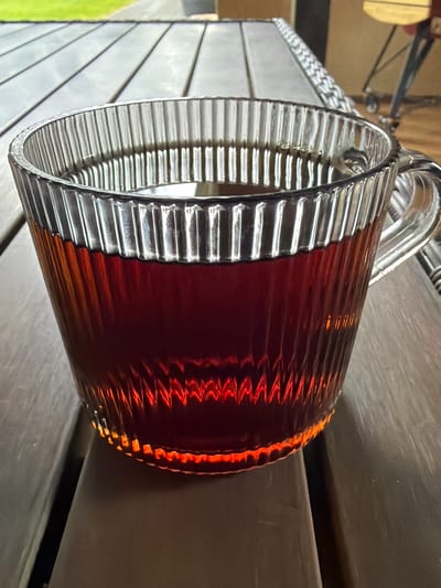 The media file is a close-up, low-angle shot of a ribbed glass pitcher filled with a dark amber liquid, possibly beer or iced tea. The pitcher is placed on a textured surface that appears to be made of weathered wooden planks. The glass has vertical ribs that create a pattern of light and shadow, with the liquid catching the light and reflecting a warm, reddish-orange glow. The handle of the pitcher is visible at the top, made of a clear, curved glass. The background is out of focus, but hints of a lighter, possibly outdoor setting can be seen, with a blur of green in the lower left corner, suggesting grass or foliage. The lighting suggests it might be daytime, with light coming from above. There is no visible text in the image.