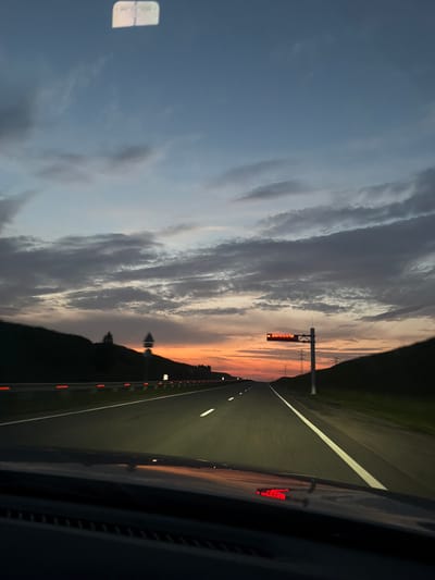 The image depicts a view from inside a vehicle on a highway at dusk. The sky is filled with dramatic clouds, with a vibrant orange and pink sunset visible on the horizon. A digital road sign is mounted on a pole, displaying red text that reads "ДОБРОГО ПУТИ" (which translates to "Have a good trip" in Russian). The highway has clear lane markings and reflective markers along the side. The foreground shows the dashboard of the vehicle, with a reflection of the red sign visible on its surface. The landscape on either side of the highway is dark and hilly. There are power lines and transmission towers visible in the distance. The scene appears to be on a highway leading out of or into the city of Luchshevo, Russia. There are no people or animals visible in the image. The overall atmosphere is one of travel and the fading light of day.