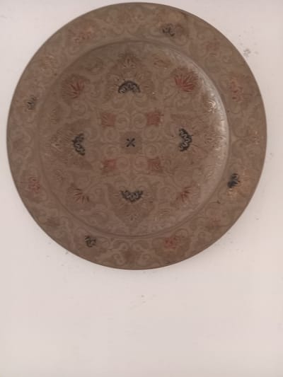 A close-up shot shows an intricately patterned, circular metal plate mounted on a white wall. The plate is predominantly a muted brown/bronze color with red and dark brown detailing within the engraved floral pattern. The lighting is soft and diffuse, resulting in minimal shadows. The wall appears to be clean and flat with a slightly textured surface. The object is likely located in Ksar-el-Kebir, Morocco. No people or actions are depicted.