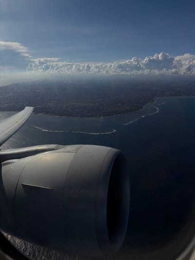 An aerial view from an airplane window shows a coastal landscape, possibly near Taoyuan City, Taiwan. The wing and engine of the aircraft are prominent in the foreground, while the land and sea are visible in the background. Clouds line the horizon, separating the land from the sky. The image is rotated.