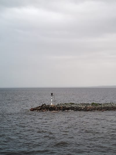 The image shows a body of water under an overcast sky. In the middle of the frame, a rocky breakwater extends into the water, topped with some vegetation. A black and white striped marker stands on the breakwater. The water is dark, and the horizon is visible in the distance. The location is Ozernoe Ust'e, Russia.