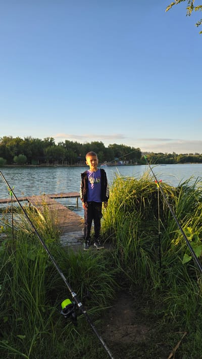 A young boy stands on a wooden dock by a lake, with fishing rods set up in the tall grass around him. He is wearing a blue t-shirt with a graphic print and dark pants. The sun is setting, casting a warm light on the scene, and the sky is a clear blue with a few wispy clouds. The lake is calm and reflects the trees on the opposite bank. There are some small buildings visible across the water. The overall impression is one of a peaceful, rural setting in the city of Қарабұлақ, Kazakhstan.
