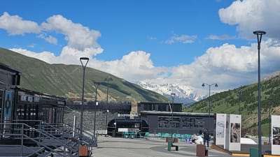 The image captures a modern commercial area situated in Kamskho, Russia, at the foot of majestic mountains. The scene is dominated by a clear blue sky adorned with fluffy white clouds. In the foreground, a paved plaza features contemporary architecture, including a black modular building with the inscription "KOFE VODA CHEKM" and a standalone "COFFEE BASE" kiosk. Several teal outdoor tables and chairs are arranged around the coffee stand. To the left, a dark gray building with metal stairs leading up to it is visible, also featuring extensive glass windows. The plaza is further furnished with modern streetlights and various display stands showcasing large-format prints.
In the mid-ground, the terrain slopes upward, transitioning from grassy hillsides to rugged, snow-capped mountains in the distance. The mountains are a prominent feature, their peaks stark against the sky. Green trees are sparsely scattered across the lower slopes of the mountains. A few people are present in the plaza; one individual is walking near the display stands, while a small group is seated at an outdoor table. The overall impression is one of a developing tourist or recreational hub in a scenic mountain environment.