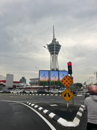 In Alor Setar, Malaysia, under a cloudy sky, a tall, distinctive observation tower stands prominently. The tower is white and features a rounded structure at its top with multiple antennas. In the foreground, a traffic light displays a red signal. Adjacent to the traffic light are two diamond-shaped yellow warning signs; one has red circles arranged in a grid, and the other displays two black arrows pointing downwards. A road with a black and white striped curb runs across the bottom of the image, with several cars parked on the side. Two large digital billboards are visible, displaying images of flowers and text that appears to say "Seni Jaya". Buildings of varying architectural styles and heights are present in the background, suggesting an urban environment. A person wearing a grey hoodie and a red helmet is partially visible on a motorcycle in the lower right corner of the image.