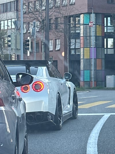 The image shows the rear of a white Nissan GT-R stopped in traffic on a street in Glattbrugg, Switzerland. The car is behind another vehicle. A pedestrian crosswalk is visible on the road with solid and dashed white lines and yellow divider lines. A building with a multi-colored window grid is in the background along with a traffic light and a street sign displaying '60', potentially indicating a speed limit. The background also includes apartment buildings with bare trees.
