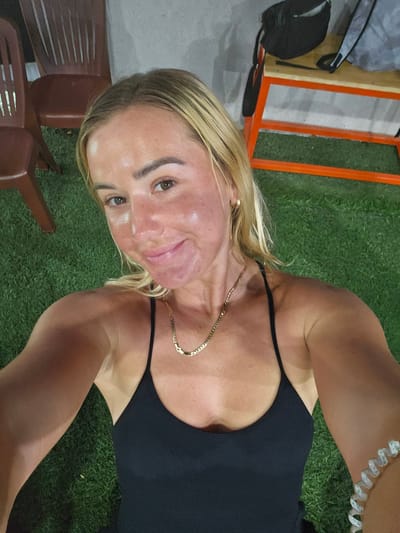 A woman with blonde hair is taking a selfie outdoors. She is wearing a black tank top and a gold chain necklace. Her skin appears red and flushed, suggesting sunburn. She is sitting on artificial turf. In the background, there are chairs and a bench with orange metal framing. The lighting suggests it is evening or nighttime. The location is Binh Thuan Ward, Vietnam.