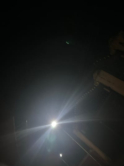 The image captures a night scene with a bright light source dominating the frame. A structure with a chain-like element is visible in the upper portion. A faint, curved object appears in the darkness to the left. The overall impression is dark and dimly lit, with the light creating stark contrast and lens flares.