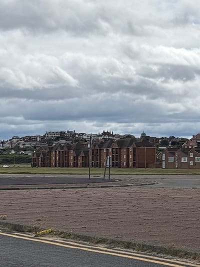 The image shows a view of the town of Wallasey, United Kingdom. In the foreground, there is a road with double yellow lines, and a rough, textured area with some grass. In the middle ground, there are several red brick apartment buildings with white-framed windows. Behind these buildings, a hill is visible, covered with a variety of houses and buildings, including some with white facades and dark roofs, and a distinctive green dome structure. The sky is overcast with heavy, grey clouds. A white car is parked on the left side of the image, near the apartment buildings. A few figures are visible walking in the grassed area between the road and the apartment buildings.