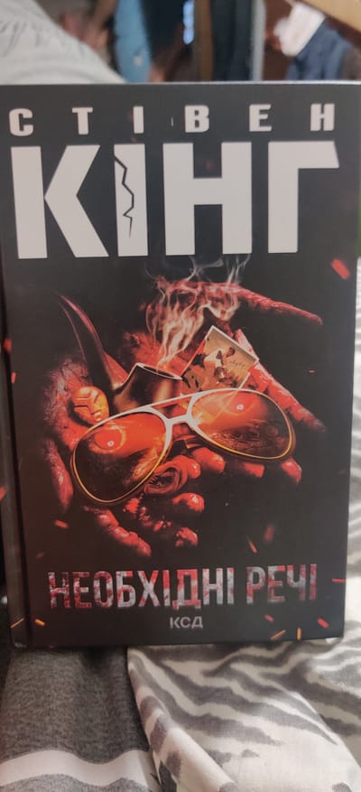 The cover of the book features a close-up of a hand, possibly male, holding a pair of sunglasses. The hand is depicted with a bloody, skeletal appearance, and flames are rising around it. The sunglasses are the focal point, with their lenses reflecting a fiery, distorted image. Behind the hand, various small, sepia-toned photographs are scattered, some showing figures in action.
Vertically along the left side of the cover, in large white capital letters, is the name "STEPHEN KING". To the right of the author's name, also running vertically, is the title of the book in a distressed, fiery font. The title appears to be in Ukrainian. The top word of the title is "НЕОБХІДНІ" and the word below it is "РЕЧІ". Beneath the title, in smaller white text, is the abbreviation "КСД".
The overall impression of the cover is dark, intense, and potentially violent, aligning with the common themes found in Stephen King's works. The color palette is dominated by dark reds, oranges, and black, with stark white text providing contrast. The background is dark and indistinct, with some small red and yellow flecks that could represent sparks or embers. The setting is not explicitly depicted beyond the imagery on the cover itself.